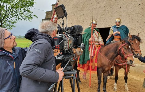 Drei Pferde mit Reitern in römischer Legionärskleidung. Davor eine große Videokamera mit Kameramann und Assistent. Im Hintergrund die Sandsteinwand eines Gebäudes