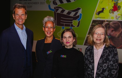 Ein Mann und drei Frauen stehen vor dem KinderKinoFest Plakat und schauen in die Kamera. 