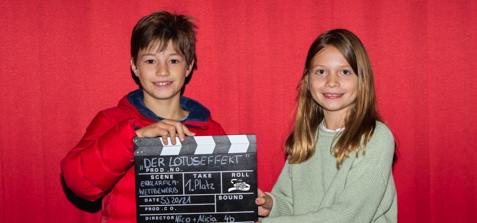 Zwei Kinder stehen vor einer roten Wand im Kino. Sie lächeln und halten eine Filmklappe in der Hand.