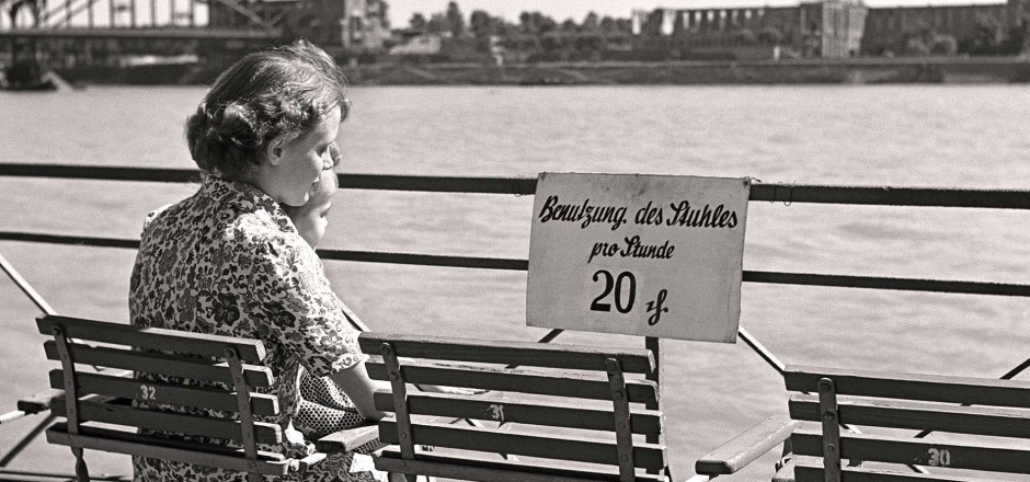 Frau sitzt mit einem Kind auf dem Schoß am Kölner Rheinufer mit Blick auf die Hohenzollernbrücke. Am Geländer ein Schild "Benutzung des Stuhls pro Stunde 20 Pfennig"