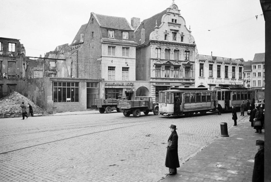 Eine Häuserzeile mit beschädigten Häusern und eine Straße mit Lastwagen und einer Straßenbahn