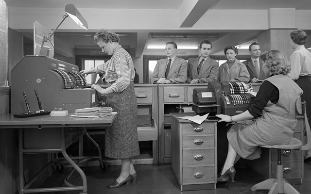 Drei Frauen arbeiten an Büromaschinen während Kunden an einer Theke warten.