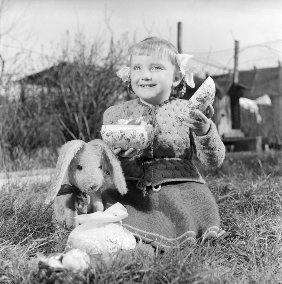 Ein Mädchen sitzt mit einem Osterei und mit einem Stofftier im Garten.