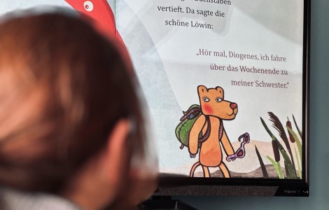 Auf der interaktiven Tafel ist ein Ausschnitt aus einem Buch zu sehen. Dort ist eine Löwin mit Rucksack illustriert. Darüber der Text: "Hör mal, Diogenes, ich fahre über das Wochenende zu meiner Schwester". 