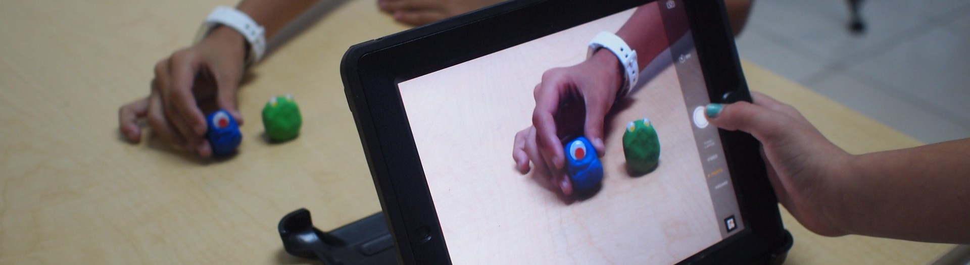 Kinderhände rücken im Hintergrund Knetfiguren zurecht, während ein zweites Paar Hände eine iPadkamera darauf ausrichten.