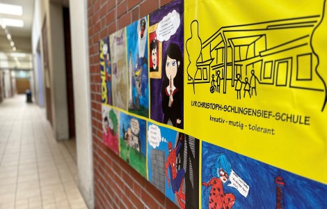 Ein Banner mit verschiedenen (Medienhelden-)Kunstwerken von Schüler*innen hängt im Schulflur, gemeinsam mit dem Slogan "LVR Christoph-Schlingensief-Schule: kreativ - mutig - tolerant"