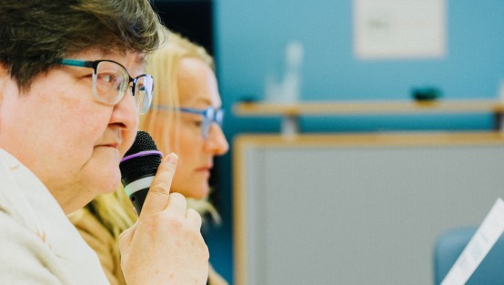 Zwei Personen sitzen an einem Tisch. Eine von ihnen hält ein Kindermikrofon in der Hand. Die andere liest etwas.