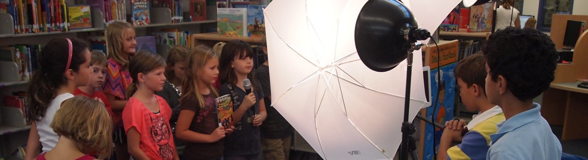  Eine Gruppe Kinder steht vor einer Bücherwand, das vorderste mit einem Mikrofon in der Hand, während weitere Kinder mit dem Rücken zur Kamera, die Szenerie mit großen Lampenschirmen ausleuchten und eine Kamera auf die Gruppe richten.