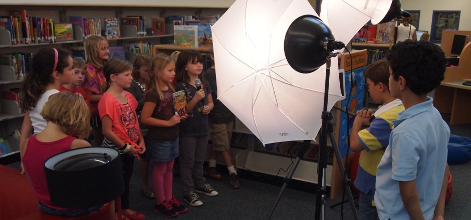  Eine Gruppe Kinder steht vor einer Bücherwand, das vorderste mit einem Mikrofon in der Hand, während weitere Kinder mit dem Rücken zur Kamera, die Szenerie mit großen Lampenschirmen ausleuchten und eine Kamera auf die Gruppe richten.