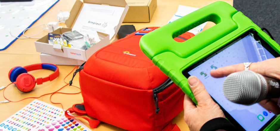 Eine Person hält ein Mikrofon und ein iPad in grüner Schutzhülle in der Hand. Im Hintergrund liegen u.a. ein orangener Rucksack und ein orangener Kopfhörer auf einem Tisch.