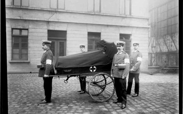 Vier Sanitäter in polizeiähnlichen Uniformen mit einer Rot Kreuz-Tragbahre auf Rädern