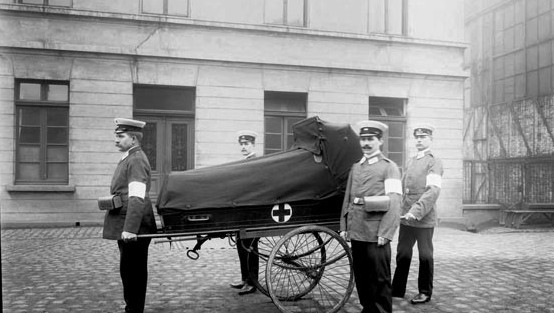 Vier Sanitäter in polizeiähnlichen Uniformen mit einer Rot Kreuz-Tragbahre auf Rädern