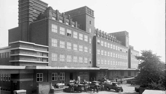 Großes Backsteingebäude mit schlichter Fassade, davor LKWs und Pferdewagen an der Laderampe