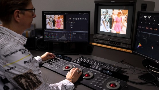 Colorgrading im Studio Auf dem Bild ist eine Person zu sehen, die vor mehreren Monitoren sitzt und professionelle Bildbearbeitungssoftware verwendet. Die Person bearbeitet ein Foto, auf dem zwei Menschen auf der Straße zu sehen sind. Einer der beiden trägt ein pinkfarbenes Kleid. Die Umgebung ist professionell eingerichtet und gut beleuchtet.