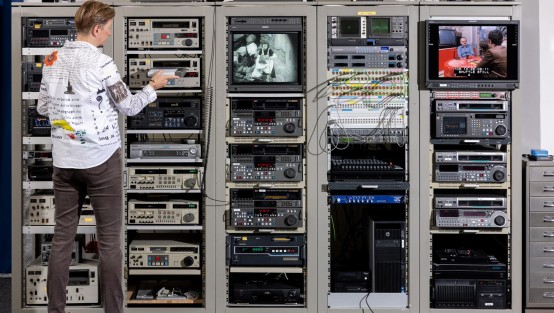Filmdigitalisierung unterschiedlichster Medien Das Bild zeigt eine Person vor einem komplexen Rack mit Audio- und Videoausrüstung. Die Person trägt ein weißes Hemd mit verschiedenen Designs und braune Hosen. Sie bedient die Geräte und überwacht zwei Monitore, die verschiedene Szenen anzeigen. Die Racks sind mit verschiedenen Geräten gefüllt, darunter Verstärker, Equalizer und andere elektronische Geräte, die mit zahlreichen Kabeln verbunden sind. Der Boden ist grau, und die Wand im Hintergrund ist hellblau.