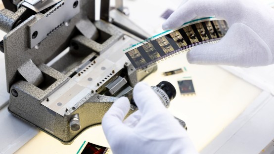 Sichtung eines Filmstreifens Eine Person in weißen Handschuhen hält einen Filmstreifen und betrachtet ihn, während sie neben einem offenen Filmprojektor steht. Verschiedene Filmstreifen und eine Filmrolle liegen auf dem Tisch.