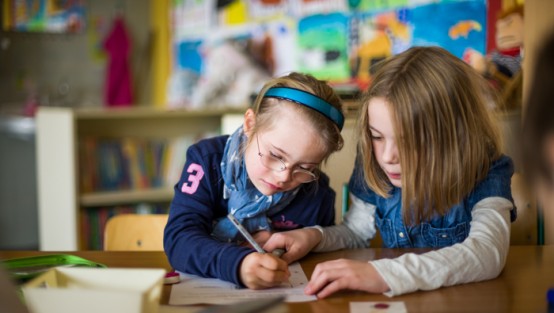 Zwei Schülerinnen, die eine schreibt etwas und die andere schaut ihr zu.
