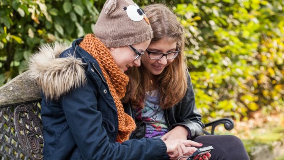 Zwei Mädchen sitzen auf einer Bank und schauen auf Ihr Handy.