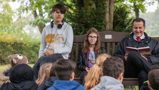 Ein Mann liest aus einem Buch und eine Schülergruppe hört ihm zu.