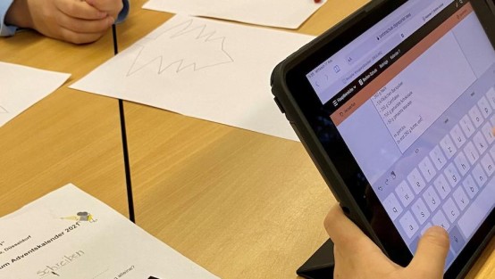Auf dem Tisch liegen Zettel, ein Kind überträgt mit dem Tablet ein Rezept auf die Website der Schulzeitung 