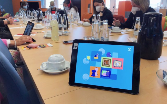 An einem gedeckten Tisch sitzen mehrere Menschen. Auf dem Tisch steht ein Tablet. Das Display leuchtet blau, die Elefanten-App ist geöffnet.