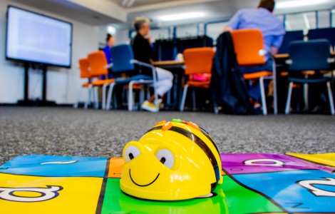 Im Vordergrund ist ein kleiner gelber Roboter mit lächelndem Gesicht (BeeBot) auf einer bunten Matte. Im Hintergrund sitzen mehrere Personen an einem Tisch.